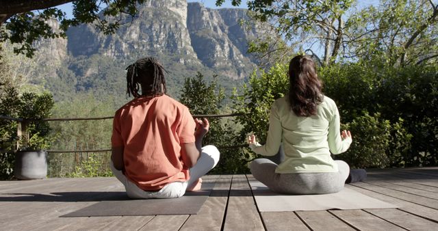 Two diverse individuals are meditating on wooden deck surrounded by nature. They are sitting cross-legged facing a scenic mountain view. The image can be used for wellness websites, yoga blogs, meditation apps, health and wellness articles. It depicts tranquility and the benefits of spending time outdoors in natural environments.