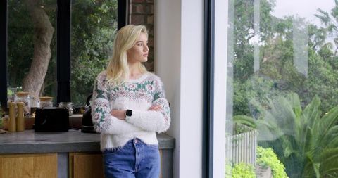 Blonde woman wearing fuzzy sweater and jeans leaning on counter gazing out window