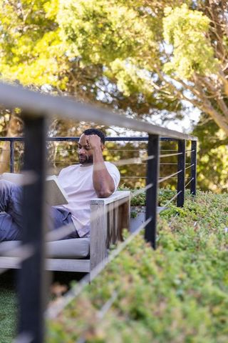 Mature african man relaxing on terrace reading book by cable railing in sunlit garden