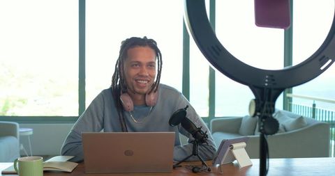 Dreadlocked content creator smiling while streaming with laptop and microphone, ring light