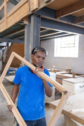 African american carpenter holding wooden frame in workshop wearing protective gear