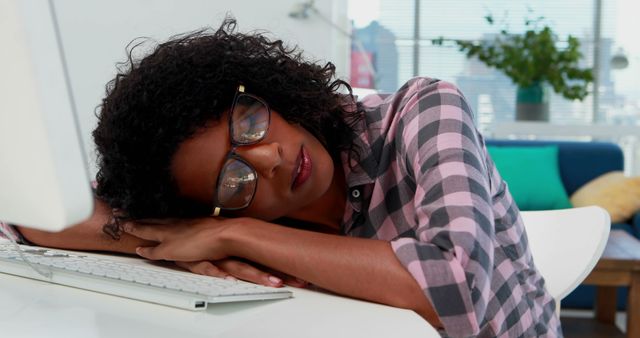 A woman with glasses and curly hair resting her head on a computer keyboard at her office desk, expressing fatigue or exhaustion. Useful for depictions of workplace stress, overworking, need for breaks, or illustrations related to health and wellness in a professional environment. Ideal for articles and campaigns about work-life balance, burnout, and the importance of rest.