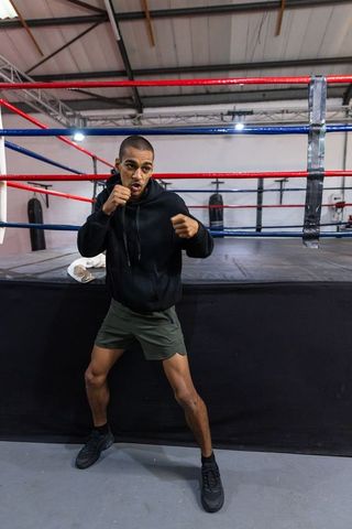 Male boxer standing on ring apron training, raising fists wearing hoodie and shorts