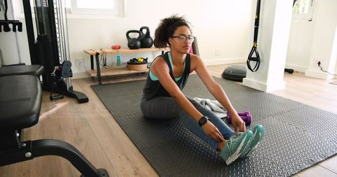 Woman stretching forward on home gym mat reaching feet with purple dumbbells and glasses