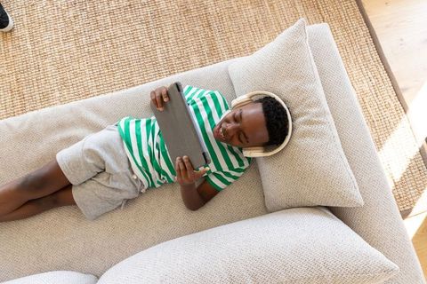 African american boy relaxing on sofa using tablet with headphones in sunlit living room