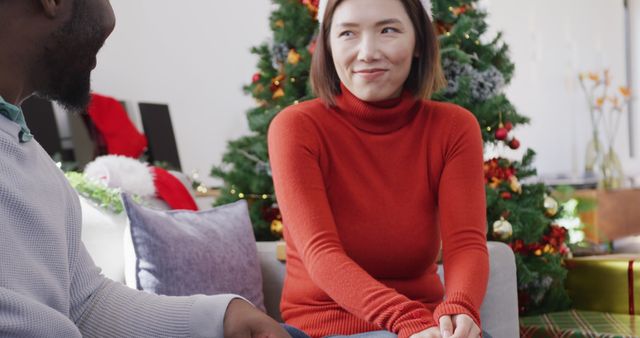 Image of happy diverse couple in santa hats exchanging christmas gift and smiling at home. Christmas, celebration, tradition, happiness and inclusivity concept.