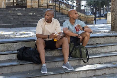 Two african men sitting on stone steps holding cold drinks and duffel bags, casual athletic friends