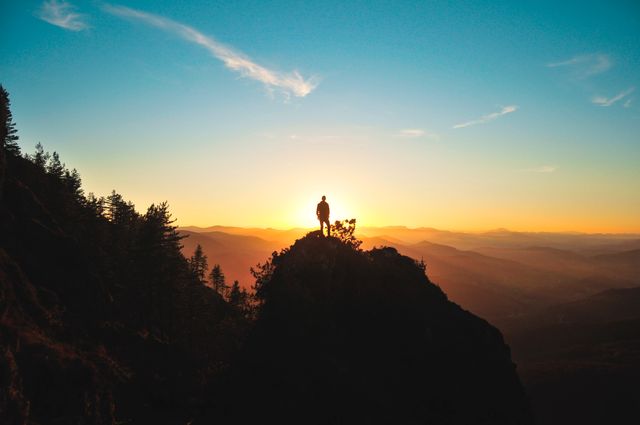 Person standing on a mountain peak at sunset, silhouetted against a vibrant, colorful sky. Ideal for use in travel and adventure marketing, inspirational posters, outdoor lifestyle blogs, or nature-related content highlighting the beauty of landscapes and the spirit of exploration and tranquility.
