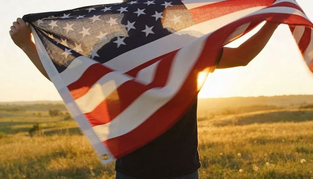 Backlit man holding and raising american flag at golden sunset over rolling field