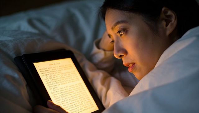 Reading backlit e-reader in bed under duvet at night, cozy quiet bedtime digital reading