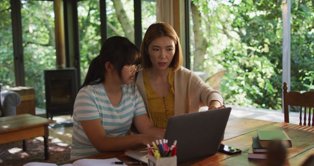 Asian mother and daughter collaborating on laptop at home in living room with big windows showcasing lush trees outside. Mother providing assistance while daughter focuses on laptop. Ideal for concepts of technology, remote education, working from home, family bonding, homeschooling, and parental support.