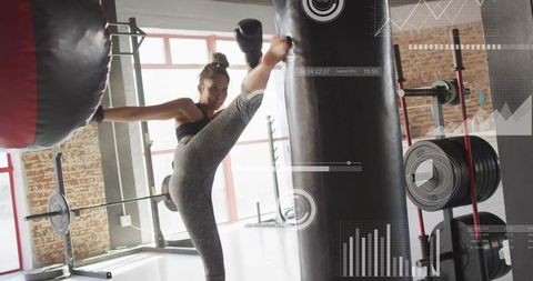 Female athlete executing high side kick striking heavy bag in gym with hud overlay