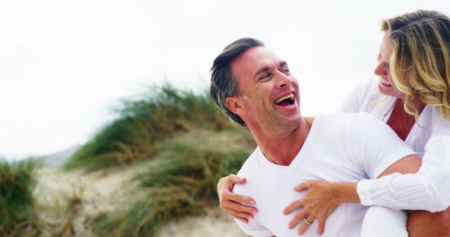 A middle-aged Caucasian man is joyfully carrying a woman on his back at a beach, both laughing and enjoying a playful moment, with copy space. Their genuine smiles and casual white attire suggest a carefree, romantic outing or vacation.