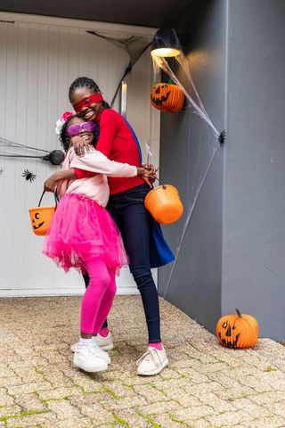African american mother and daughter hugging on porch in halloween costumes with jack-o-lanterns