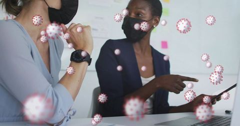 Masked colleagues collaborating and pointing at tablet with virus overlay in office meeting