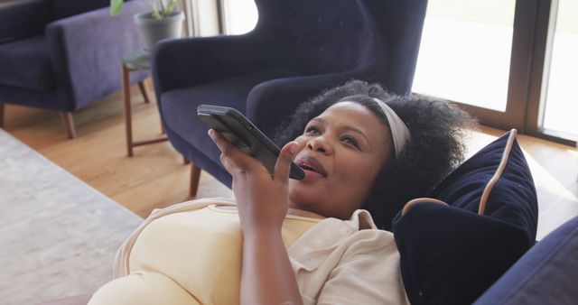 Pregnant woman is lying on a couch comfortably while using a smartphone for voice control. Perfect for content related to maternity, modern lifestyles, smart home technology, or relaxation. Can be used in articles, blogs, social media posts, or advertisements focusing on motherhood, smart devices, home comfort, or lifestyle topics.