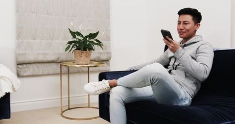 Young Asian Man Relaxing on Elegant Navy Velvet Couch with Smartphone