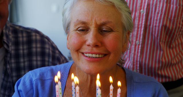 A senior Caucasian woman smiles joyfully in front of a birthday cake lit with candles, with copy space. Her expression captures a moment of happiness and celebration during a special occasion.