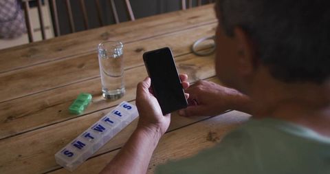 Senior man holding smartphone and checking weekly pill organizer on wooden table