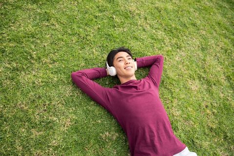 Asian young man relaxing on grass listening to music with white headphones maroon shirt