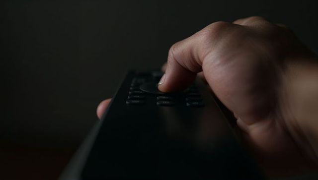 Thumb pressing TV remote control in low-light room, moody close-up for entertainment