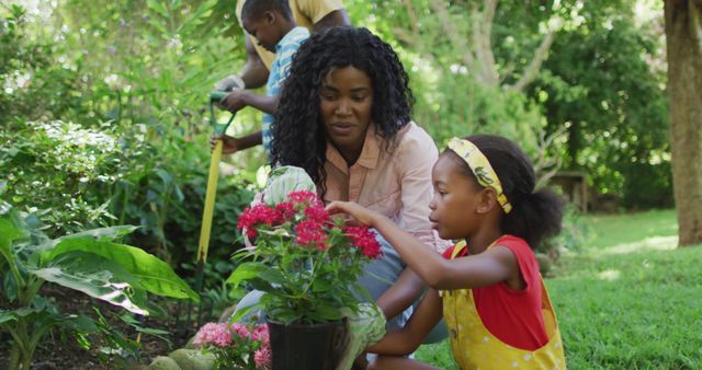 African American family enjoying a sunny day while gardening outdoors. A woman and child focus on planting flowers together while a man and another child work in the background. Ideal for themes of family bonding, outdoor activities, nature, gardening, and teamwork. Suitable for promoting family values, gardening products, and outdoor lifestyle campaigns.