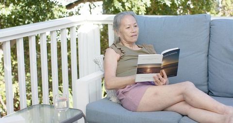 Senior asian woman reading paperback on sunlit deck sofa, relaxing on patio with glass of water
