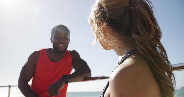 Friends are enjoying a casual conversation while standing near railing at a beach. One person wears a red sleeveless shirt, and the other has their hair in a ponytail. The scene highlights a relaxed, friendly atmosphere and can be used to promote outdoor activities, summer events, or lifestyle content.