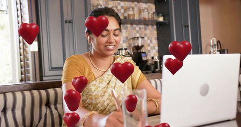 Indian woman in saree writing on laptop, smiling in kitchen nook with floating hearts