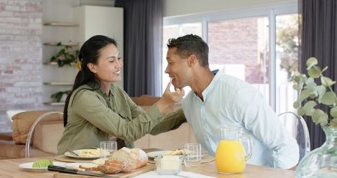 Asian woman feeding partner breakfast at kitchen table, sharing playful morning moment