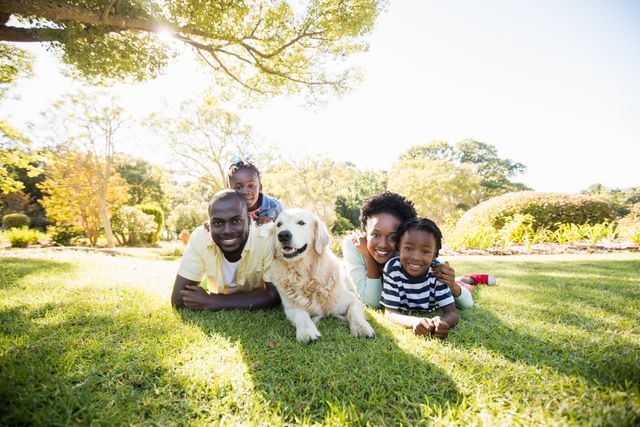 This image shows a joyful African American family lying on the grass in a park with their golden retriever. The parents and two children are smiling and enjoying a sunny day outdoors. Perfect for use in advertisements, family-oriented content, pet care promotions, and articles about family bonding and outdoor activities.