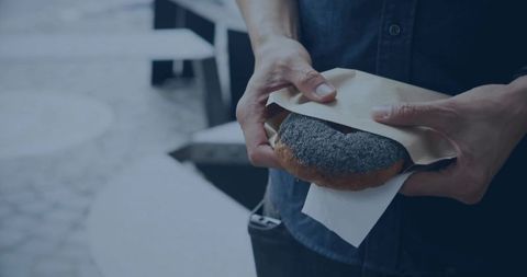 Mid-adult man pulling paper wrapper from seeded donut while standing at urban cafe plaza