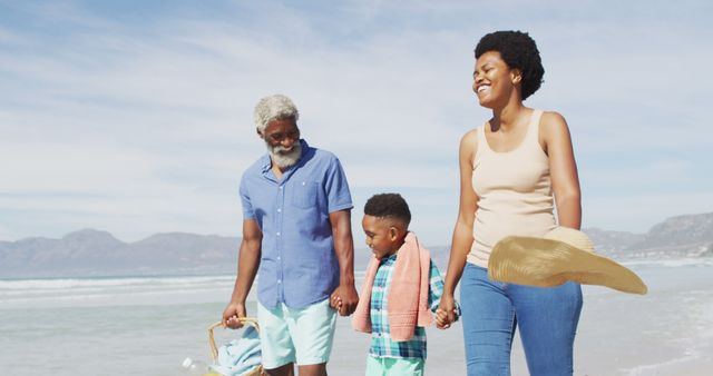African American family enjoying sunny beach day. Grandparent, mother, and child walking along shoreline, carrying beach essentials. Perfect for topics on family bonding, leisure, vacations, and multi-generational activities.
