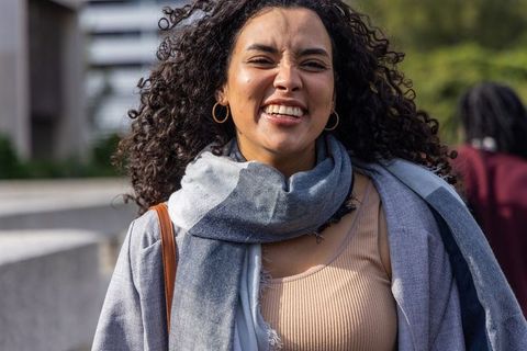 Joyful curly-haired woman walking across urban bridge wearing grey scarf and coat