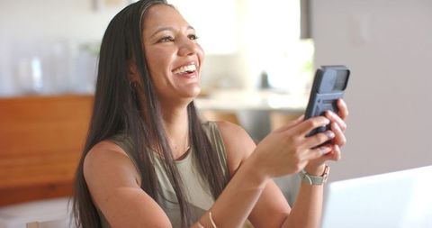 Laughing woman holding flip phone video calling at bright kitchen table with laptop