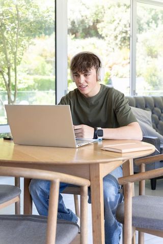 Teenage male studying at wooden dining table with headphones and laptop in sunlit home
