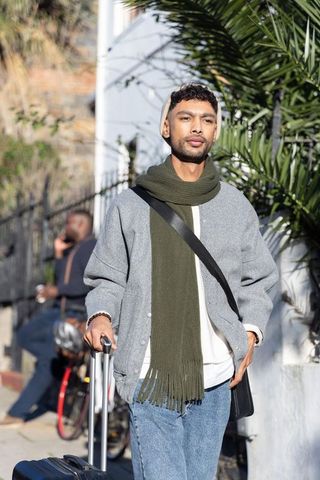 Sunlit urban traveler walking sidewalk pulling rolling suitcase wearing beanie and scarf