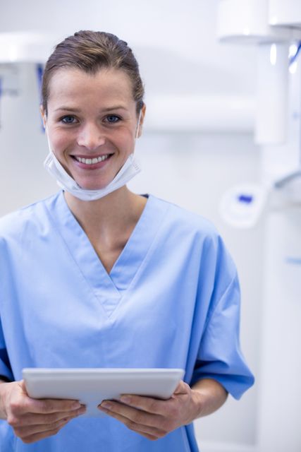 Smiling dental assistant holding digital tablet in dental clinic, wearing blue uniform and standing in a modern dental setting. Suitable for use in healthcare, dental, and medical technology content to depict professional and friendly staff in a clinical environment.
