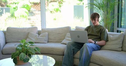 Teen sitting on sofa using laptop and headphones wearing smartwatch in bright living room