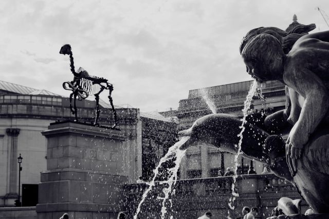 Black and white image capturing classical sculptures and a striking skeletal animal statue above a fountain. Water sprays dramatically from the structure, creating a dynamic effect. Suitable for use in travel articles, historical features, or architecture studies.