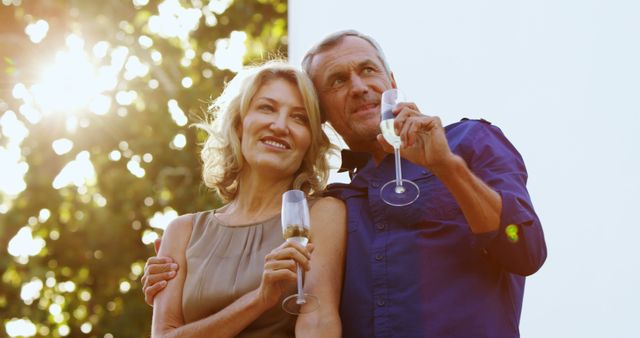 A middle-aged Caucasian couple enjoys a romantic moment with glasses of champagne, with copy space. Their content expressions and the outdoor setting suggest a celebration or a special occasion.