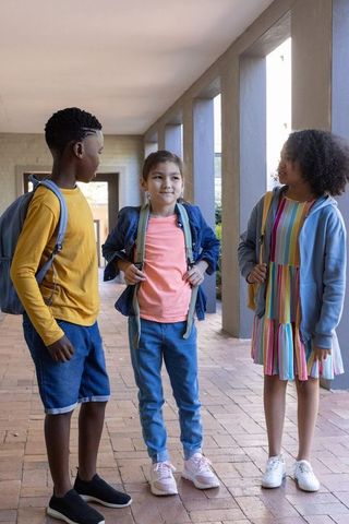 Three diverse schoolchildren standing and talking in corridor with backpacks and denim jacket