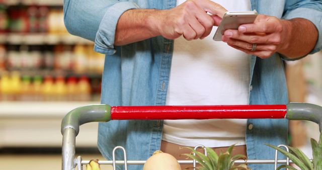 Man using smartphone while grocery shopping at the supermarket. Visible fruits and vegetables in shopping cart. Ideal for topics related to online grocery shopping, modern retail experience, or in-store technology integration.