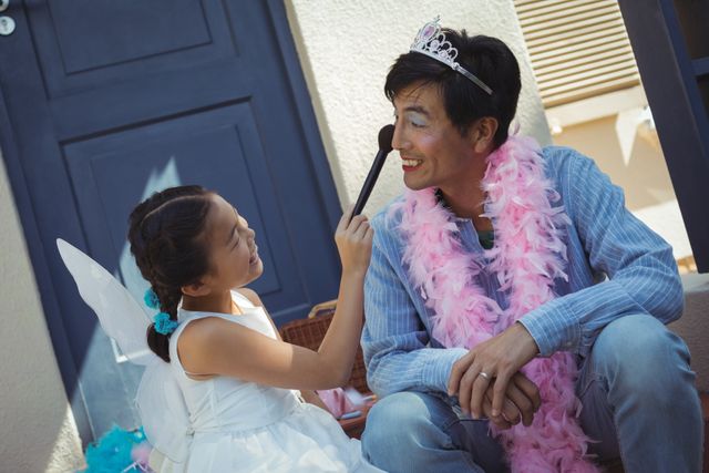 Young girl in fairy costume applying makeup to her father's face, both smiling and enjoying the moment. Perfect for themes related to family bonding, playful activities, childhood imagination, and parenting. Ideal for use in family-oriented advertisements, parenting blogs, and social media posts celebrating father-daughter relationships.
