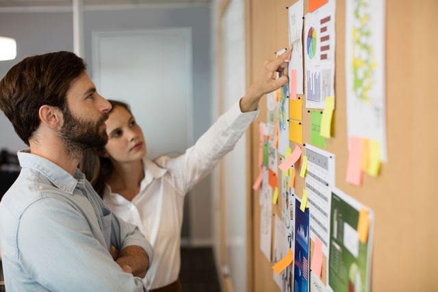 Young businessman and female colleague analyzing charts and graphs on a soft board in an office. Ideal for use in business presentations, teamwork and collaboration concepts, project planning, and corporate strategy discussions.