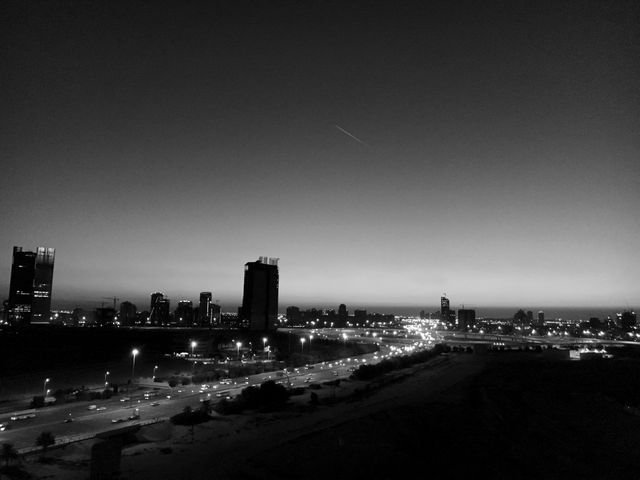 Dramatic black and white view of a city's skyline at dusk. Highway filled with lights as cars commute during the evening. Silhouetted buildings create a striking urban landscape against a darkening sky. Can be used for urban development, transportation themes, modern architectural designs, or nightlife concepts.