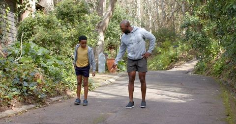 African american dad walking with son on tree-lined lane pointing at pavement for bonding