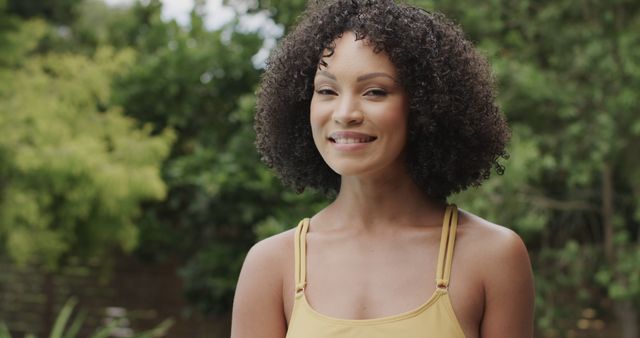 A joyful young woman with curly hair is smiling outdoors, surrounded by lush greenery. She is wearing casual clothing and appears relaxed, embodying a lifestyle of healthy living and happiness. Ideal for use in promotional materials for wellness brands, lifestyle blogs, nature retreats, or advertisements focusing on fitness and well-being.