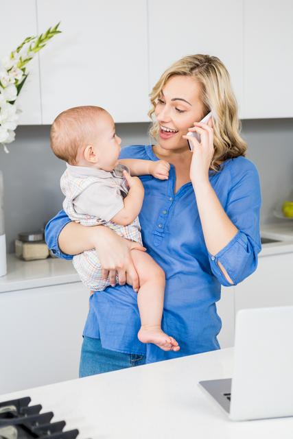 Mother holding baby while talking on mobile phone in kitchen. Ideal for content related to parenting, family life, multitasking, modern motherhood, home lifestyle, and communication technology.