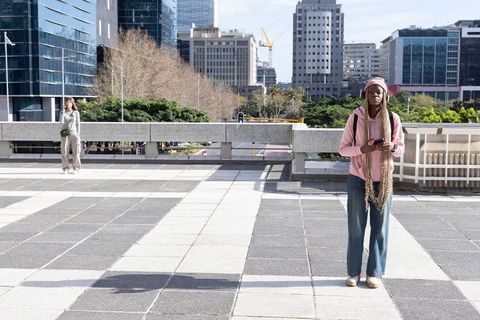 African american woman standing on city terrace holding smartphone and backpack listening to music